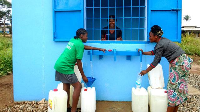 Two people filling jerrycans with water at a watering station with a smiling station attendant inside a booth